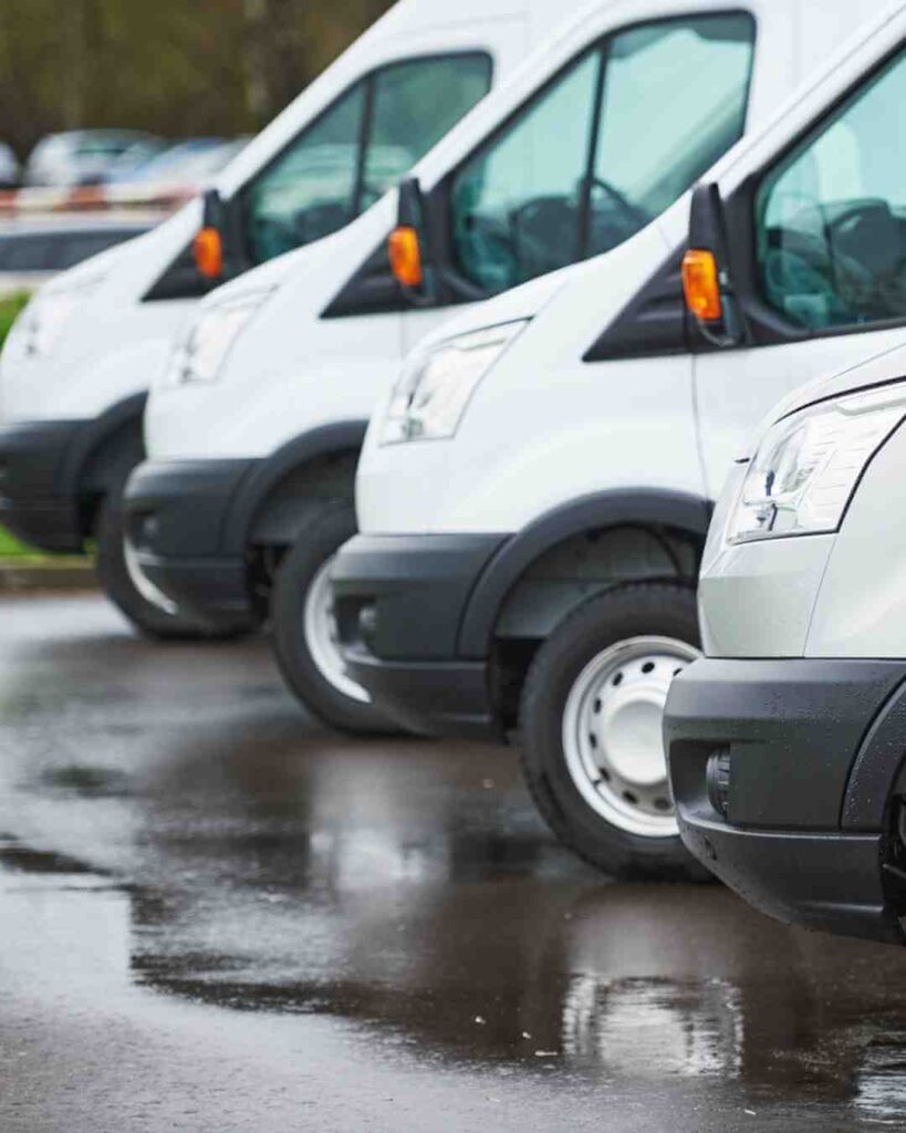 Line of white commercial vans parked on wet pavement for insurance fleet coverage.