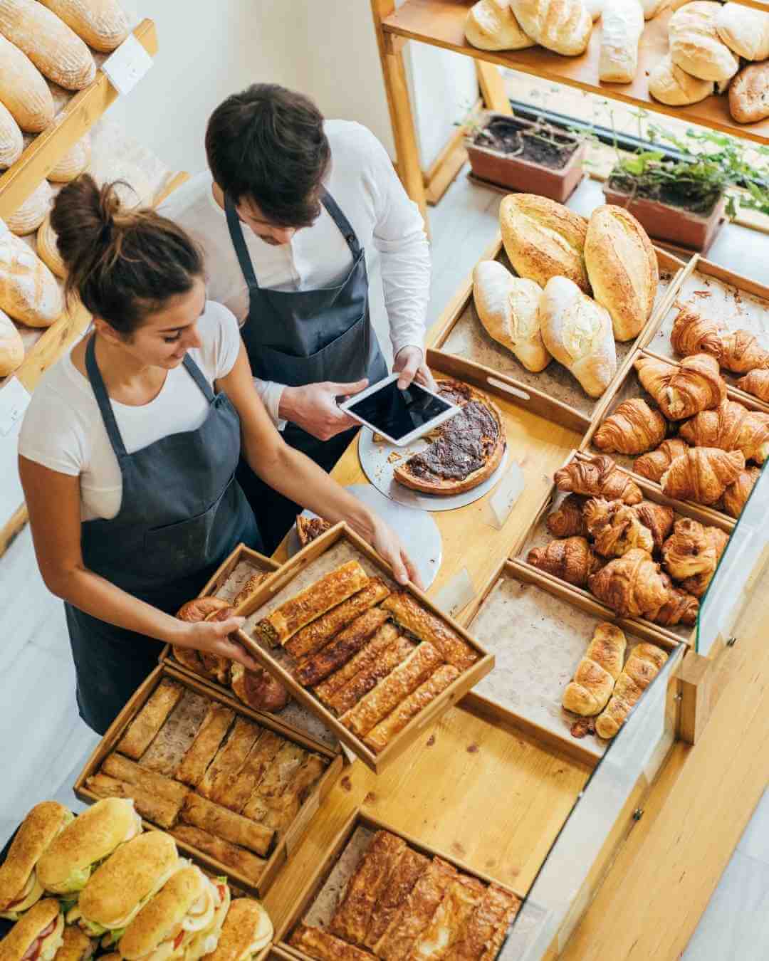 Two bakers in aprons arranging baked goods in a bakery shop.